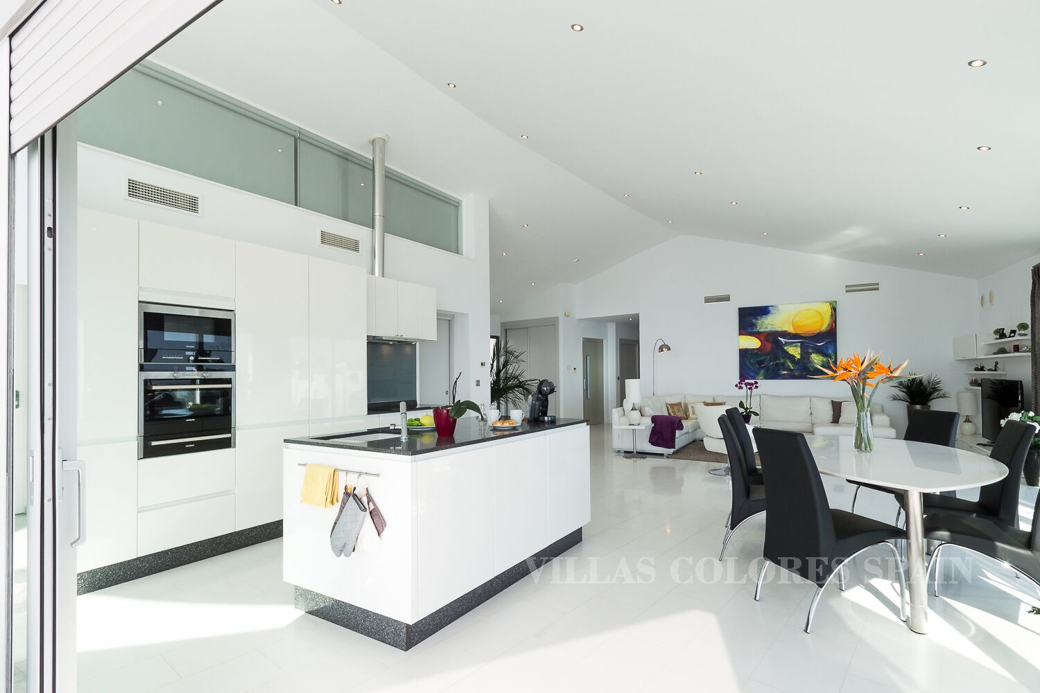 A modern kitchen with built-in appliances, a dining area with a round table and chairs, and a living space with a sofa and a large painting.