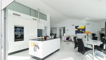 A modern kitchen with built-in appliances, a dining area with a round table and chairs, and a living space with a sofa and a large painting.