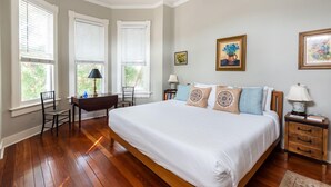 A bedroom with a large bed, wooden floor, two windows with blinds, a desk, and a chair.