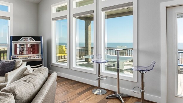 Een woonkamer met een bank, twee barkrukken en grote ramen met uitzicht op een balkon met buitenmeubilair.