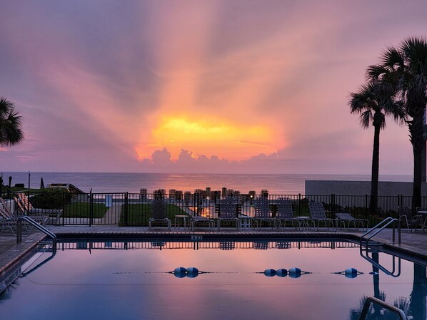 A swimming pool with lounge chairs and a view of the sunset over the ocean.