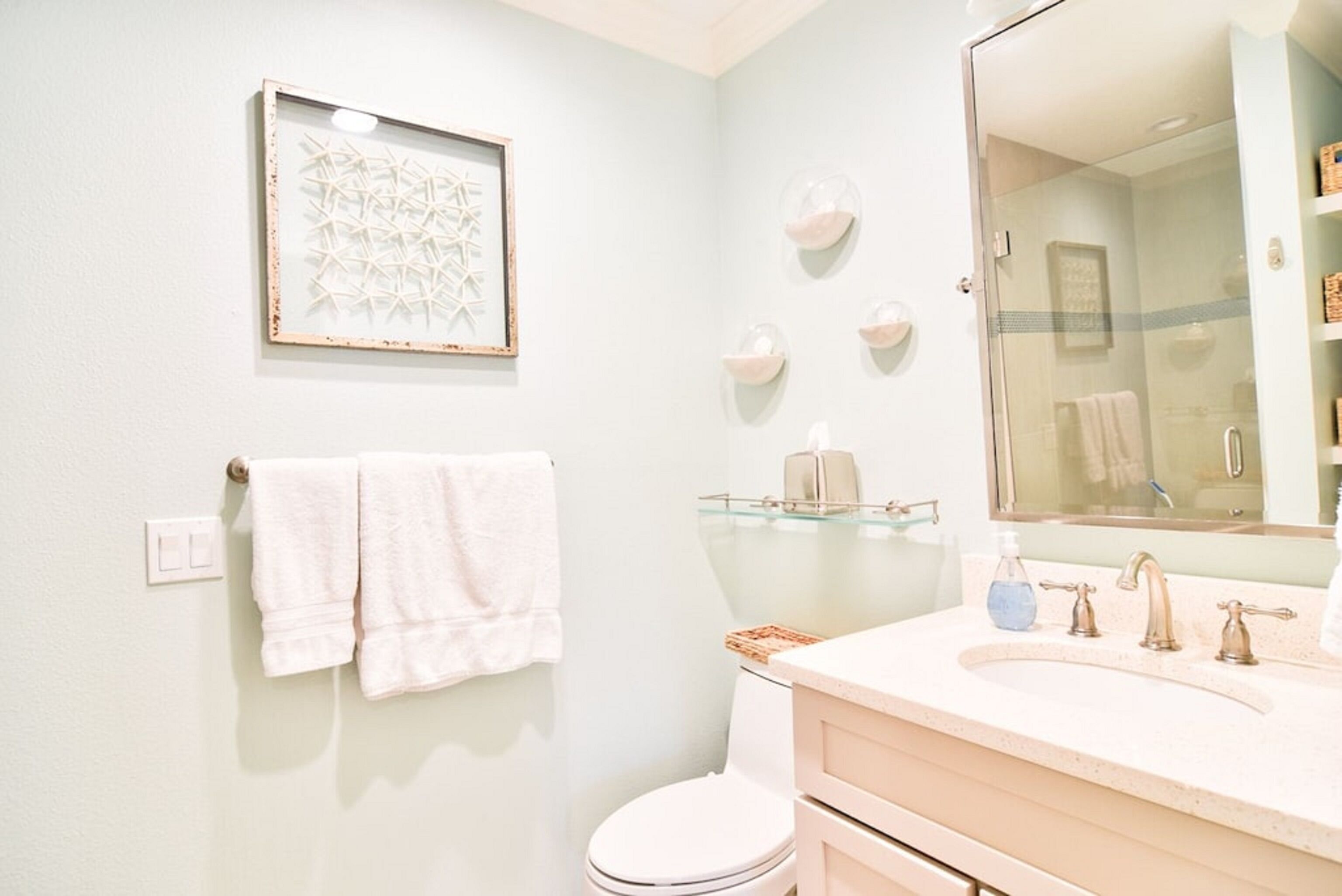 A bathroom with a toilet, sink, and mirror. There are towels hanging on the wall and a framed picture above the toilet.