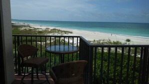 A balcony with wicker chairs and a glass table overlooking a beach with clear turquoise water and white sand. - Anna Maria Island Getaway (Holmes Beach)