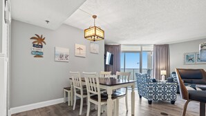 A dining area with a wooden table and chairs, a patterned sofa, and a TV mounted on the wall. - Oceanfront family fun, multiple pools, great views + Free Attraction Tickets! (Myrtle Beach)