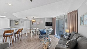 A modern living room with a sofa, a glass coffee table, and a dining area with chairs and a table. - Oceanfront family fun, multiple pools, great views + Free Attraction Tickets! (Myrtle Beach)