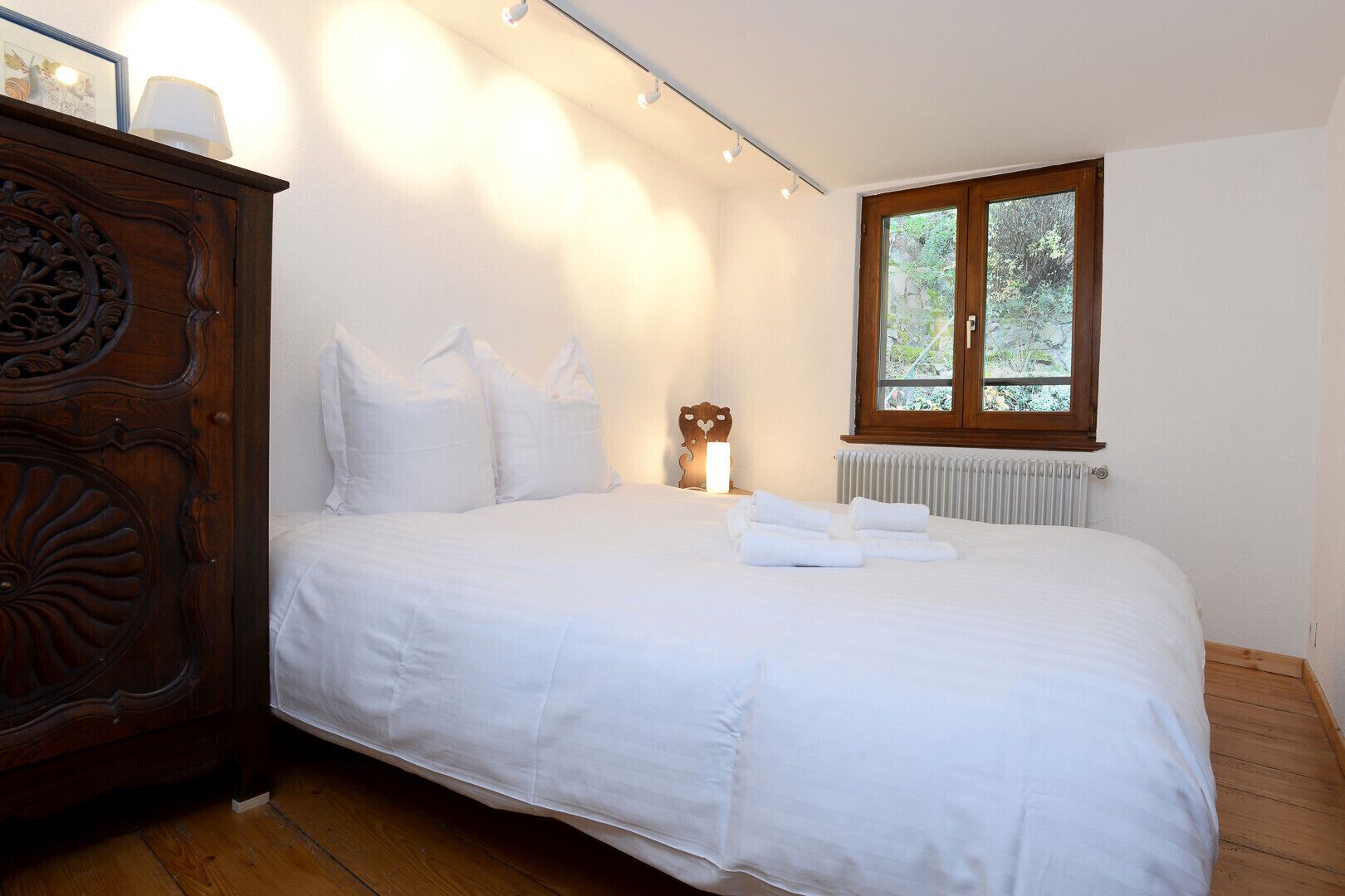 A bedroom with a large bed, a wooden dresser, a radiator, and a window with a view of trees.