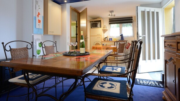 Un coin repas avec une table en bois, des chaises en osier et un coussin bleu orné d’un motif de bouée de sauvetage. Une bouteille de vin est posée sur la table.