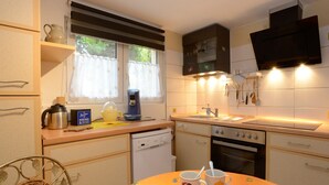 Une cuisine compacte équipée d’un évier, d’une cuisinière et d’un four à micro-ondes. On y trouve une fenêtre avec des rideaux, une chaise et une table avec des tasses.
