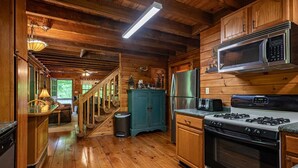A wooden cabin interior with a kitchen featuring a stove, microwave, refrigerator, and a staircase leading to a lofted area.