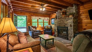 A cozy cabin interior with a stone fireplace, wooden furniture, and a large window with curtains.