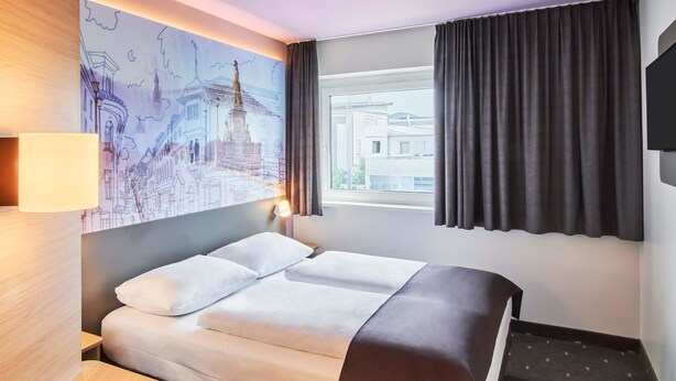 Habitación de hotel con cama, escritorio, lámpara, cuadro de un paisaje urbano, ventana con cortinas y televisor en la pared.