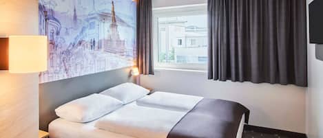 Habitación de hotel con cama, escritorio, lámpara, cuadro de un paisaje urbano, ventana con cortinas y televisor en la pared.