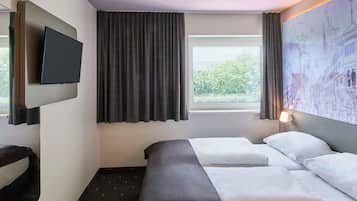 A hotel room with a bed, a flat-screen TV mounted on the wall, a window with curtains, and a wall-mounted lamp.