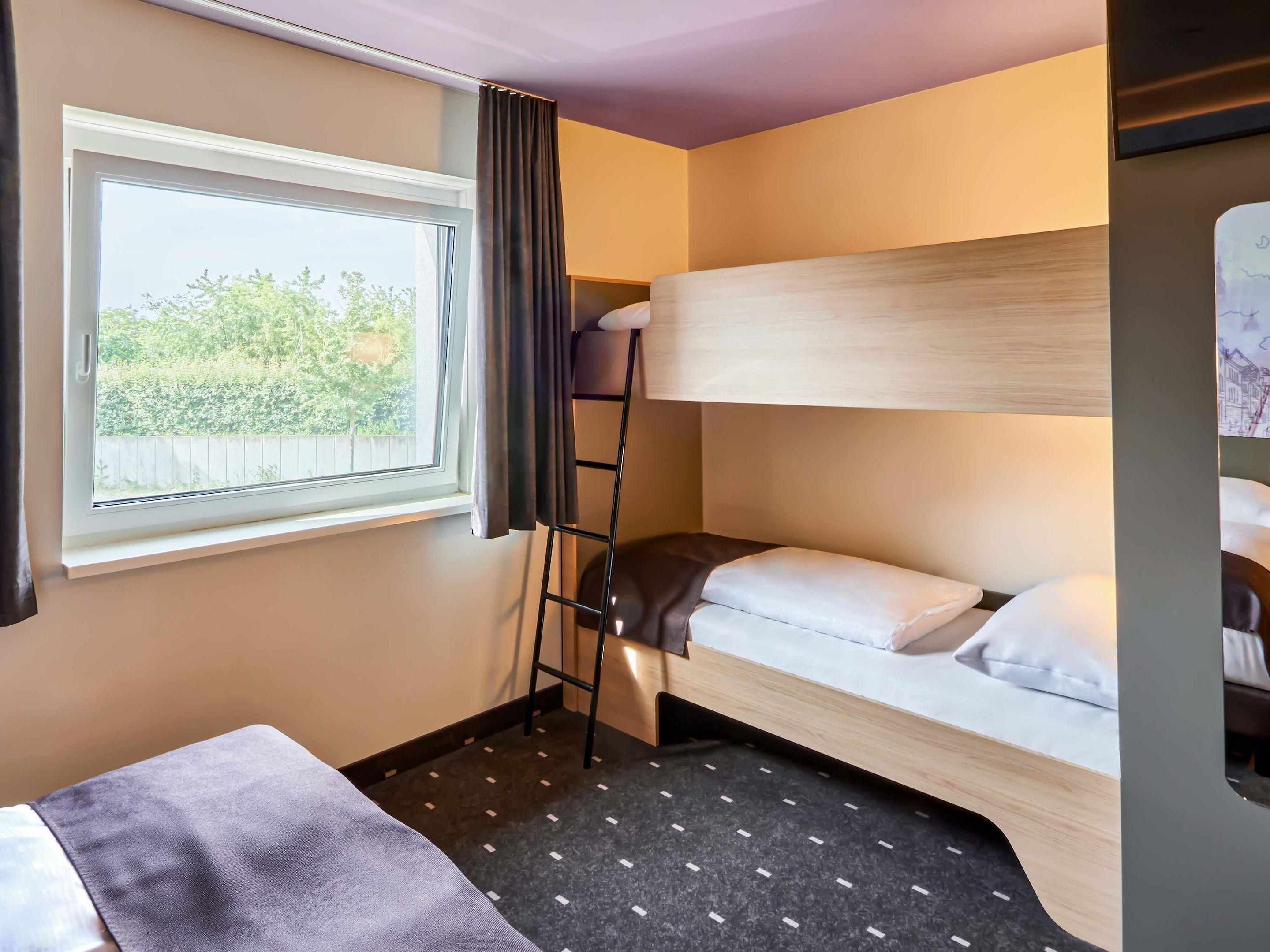 Habitación de hotel compacta con literas, una ventana pequeña con vistas a zonas verdes, una mesita de noche y un suelo estampado.