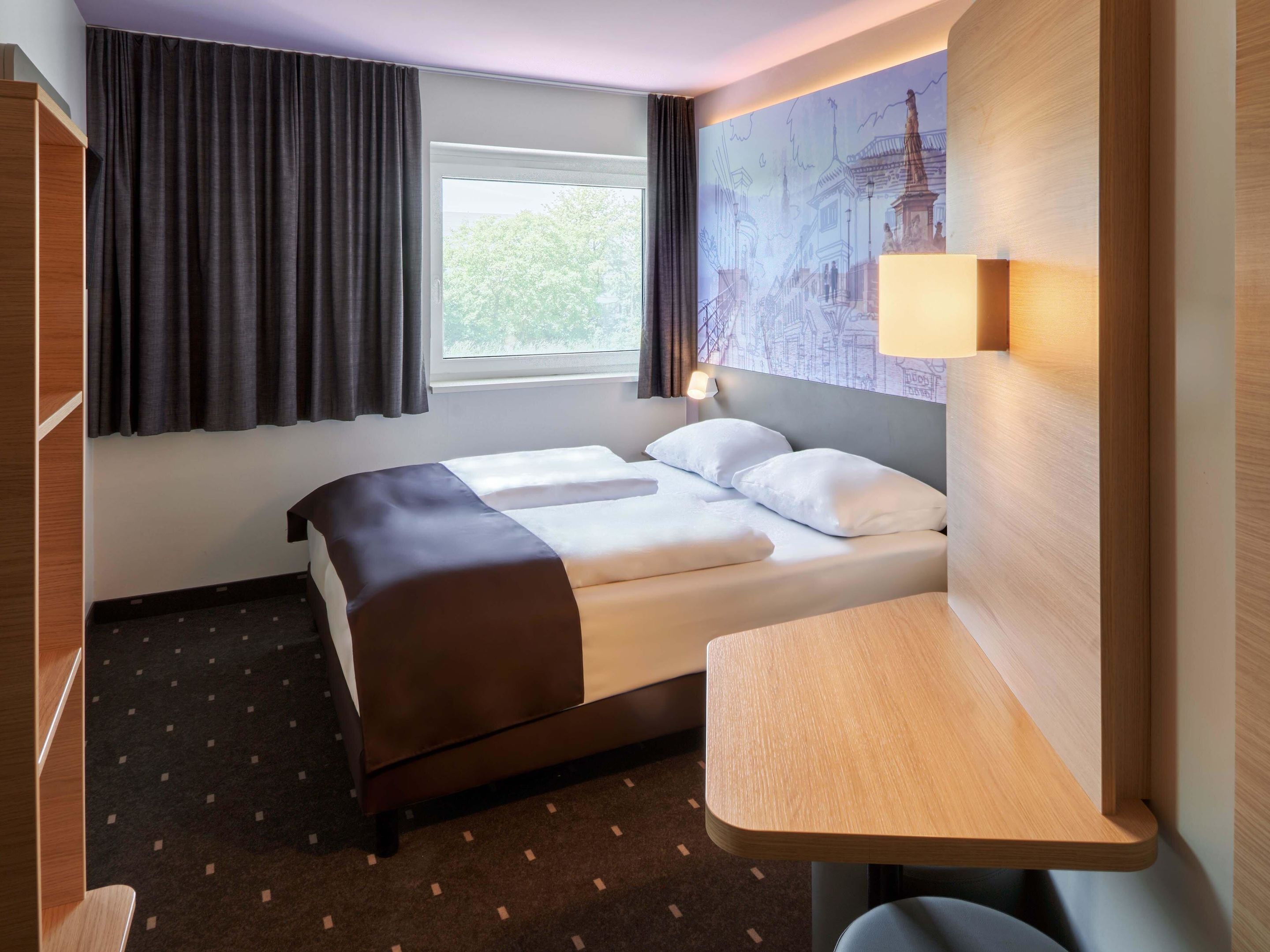 Een hotelkamer met een bed, een bureau, een raam met gordijnen en een muurschildering van een stadsgezicht.