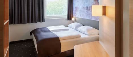 A hotel room with a bed, a desk, a window with curtains, and a wall with a cityscape mural.