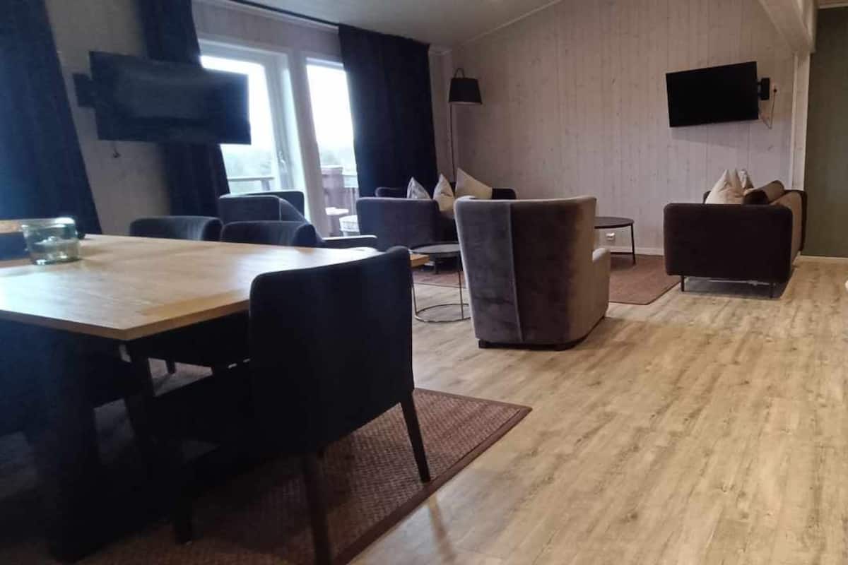 A room with a wooden floor, a dining table with chairs, a sofa, a bed, and a TV mounted on the wall.