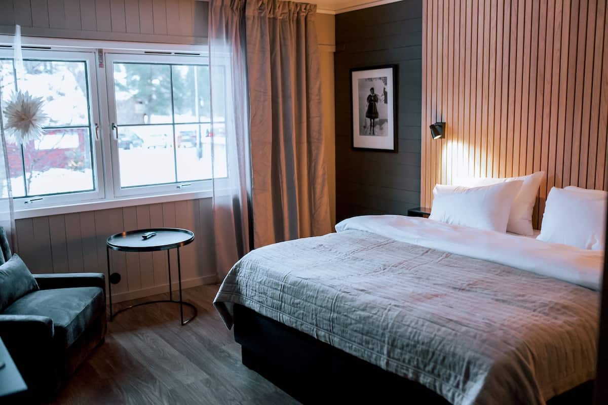 A hotel room with a bed, a chair, a small table, a window with curtains, and a framed picture on the wall.