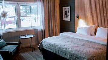 A hotel room with a bed, a chair, a small table, a window with curtains, and a framed picture on the wall.