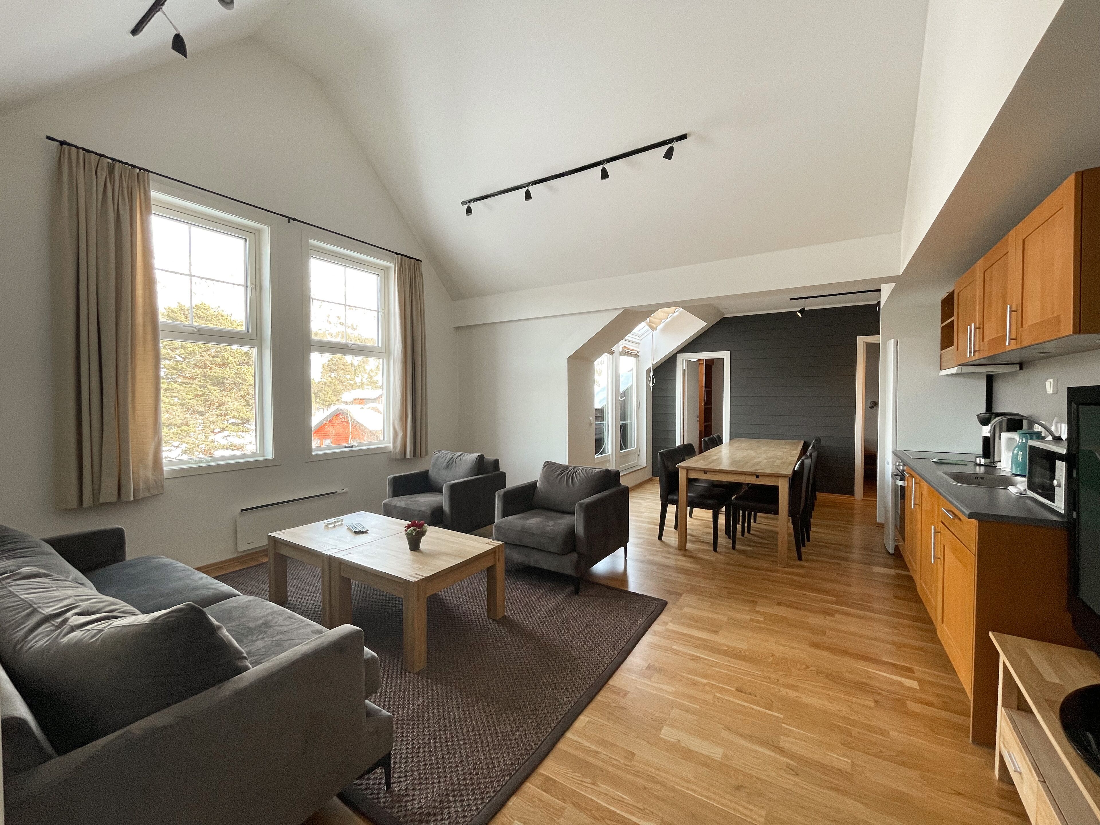 Una sala de estar moderna con un sofá, sillones, una mesa de centro y una mesa de comedor. Hay una zona de cocina con armarios y electrodomésticos. Amplias ventanas ofrecen luz natural y vistas al exterior.