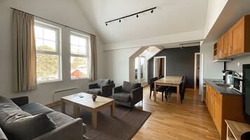 Una sala de estar moderna con un sofá, sillones, una mesa de centro y una mesa de comedor. Hay una zona de cocina con armarios y electrodomésticos. Amplias ventanas ofrecen luz natural y vistas al exterior.