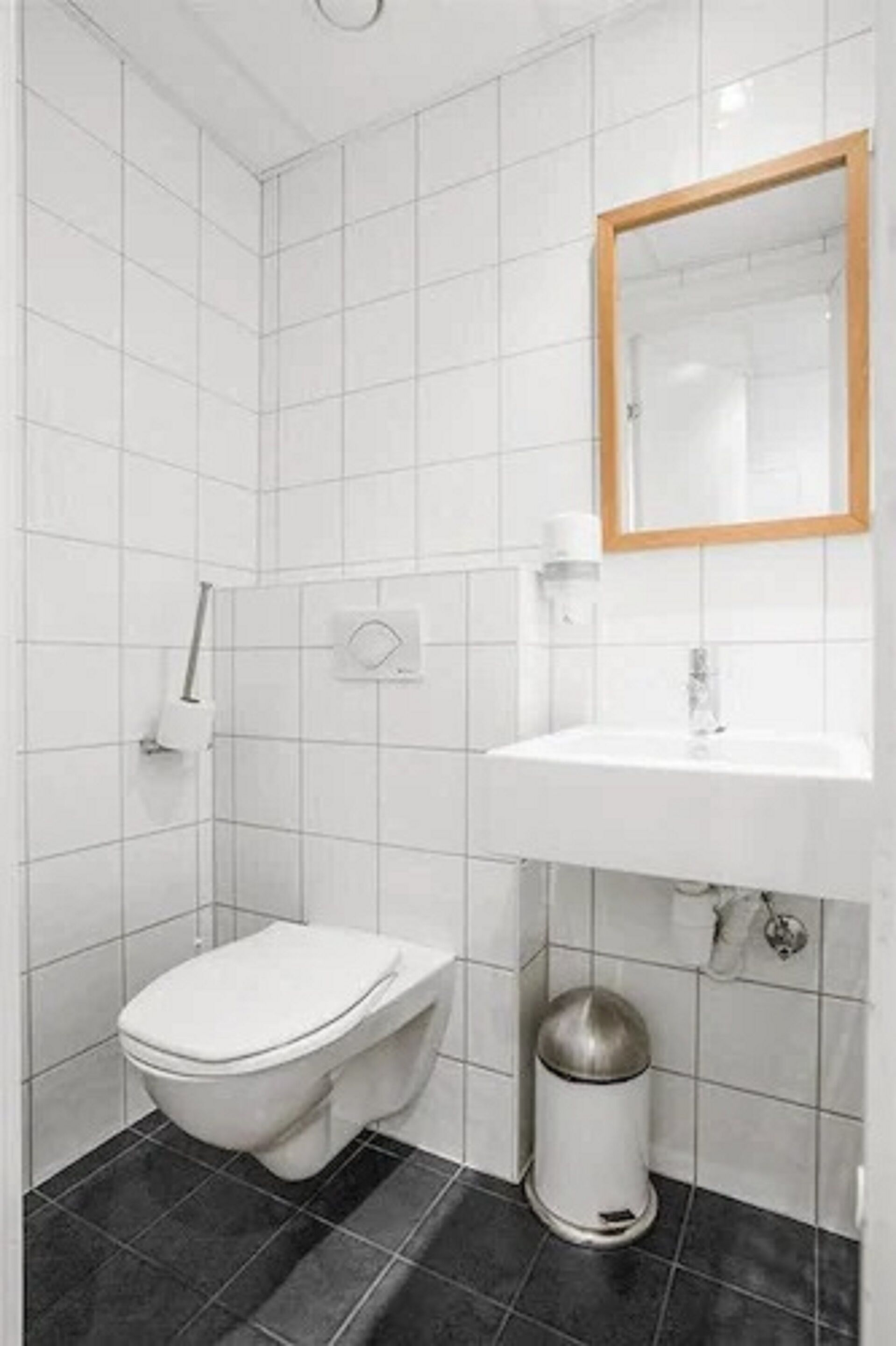 Ein kleines, modernes Badezimmer mit weißen Fliesen, einer weißen Toilette, einem Waschbecken inklusive Spiegel und einer Mülleimer.