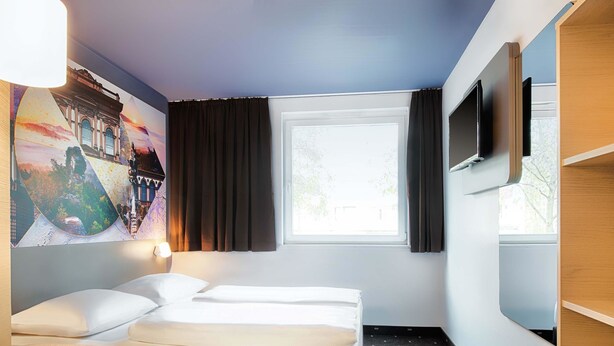 A hotel room with a bed, a desk, a chair, a TV, and a window with curtains. - B&B Hotel Kaiserslautern (Kaiserslautern)