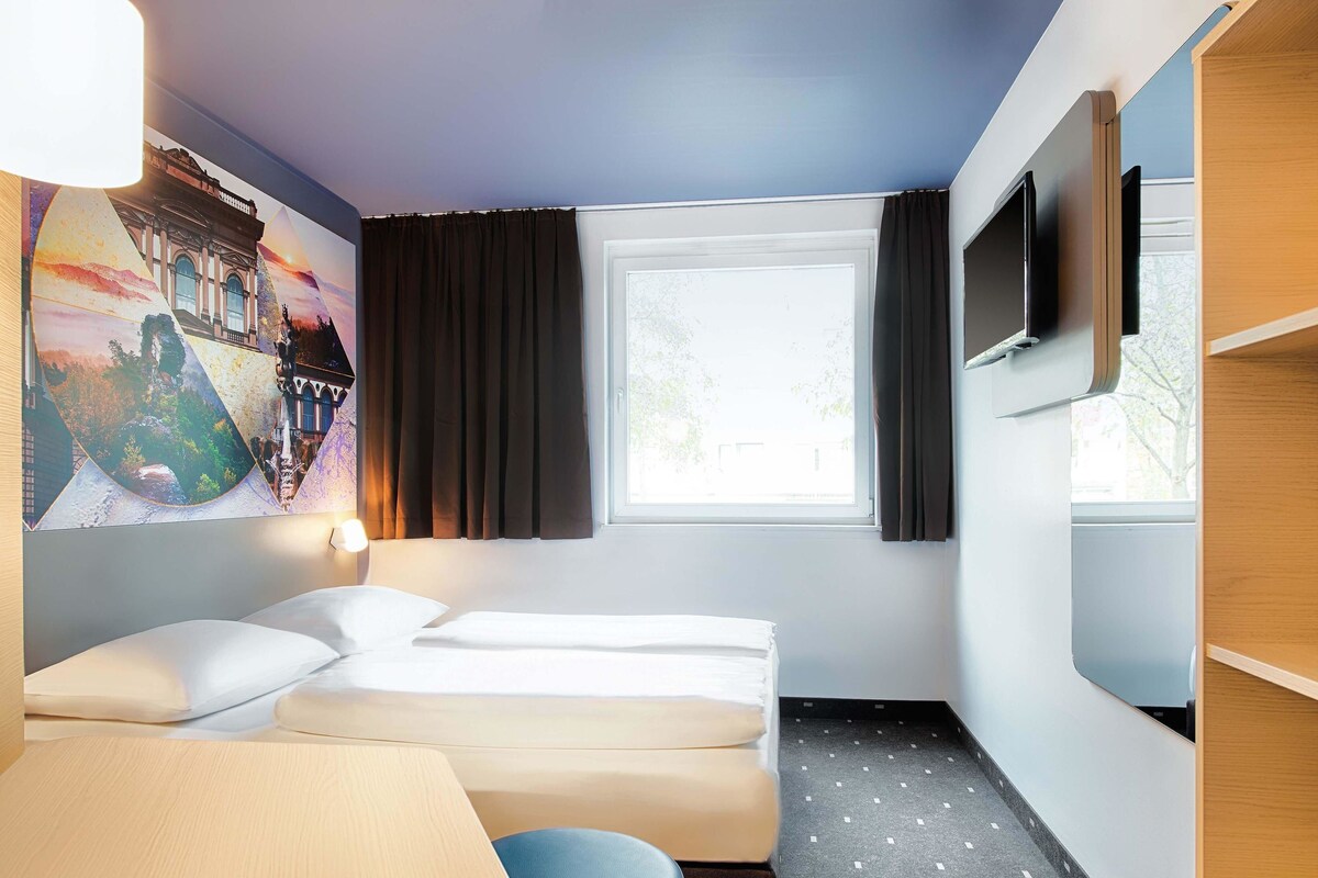A hotel room with a bed, a desk, a chair, a TV, and a window with curtains.