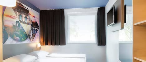 Una habitación de hotel con cama, escritorio, silla, televisor y ventana con cortinas.