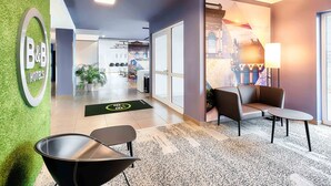 A hotel lobby with a green wall, a reception desk, seating area, and a view of the cityscape through the windows. - B&B Hotel Kaiserslautern (Kaiserslautern)