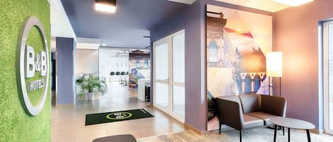 A hotel lobby with a green wall, a reception desk, seating area, and a view of the cityscape through the windows.