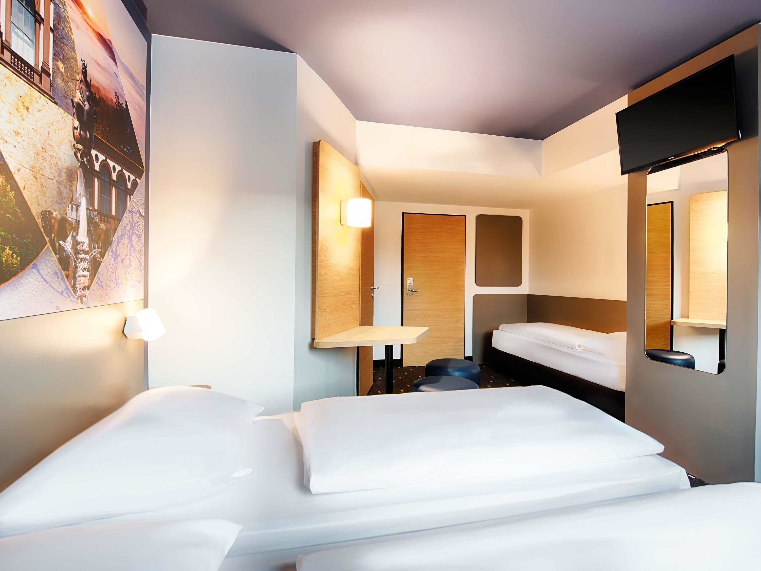 Ein Hotelzimmer mit zwei Betten, einem kleinen Tisch, einem Spiegel und einem am Wand montierten Fernseher.