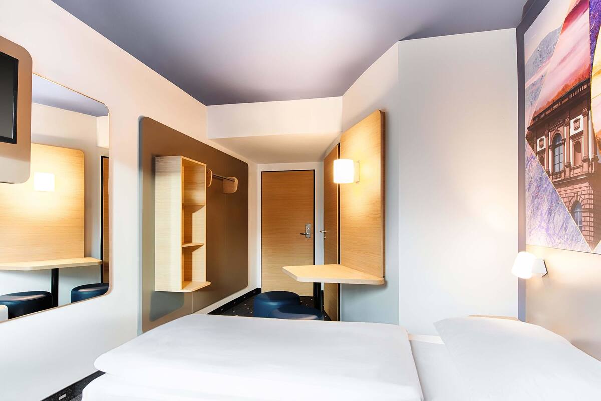 A modern hotel room with a bed, a mirror, a wooden shelf, and a wall-mounted light.