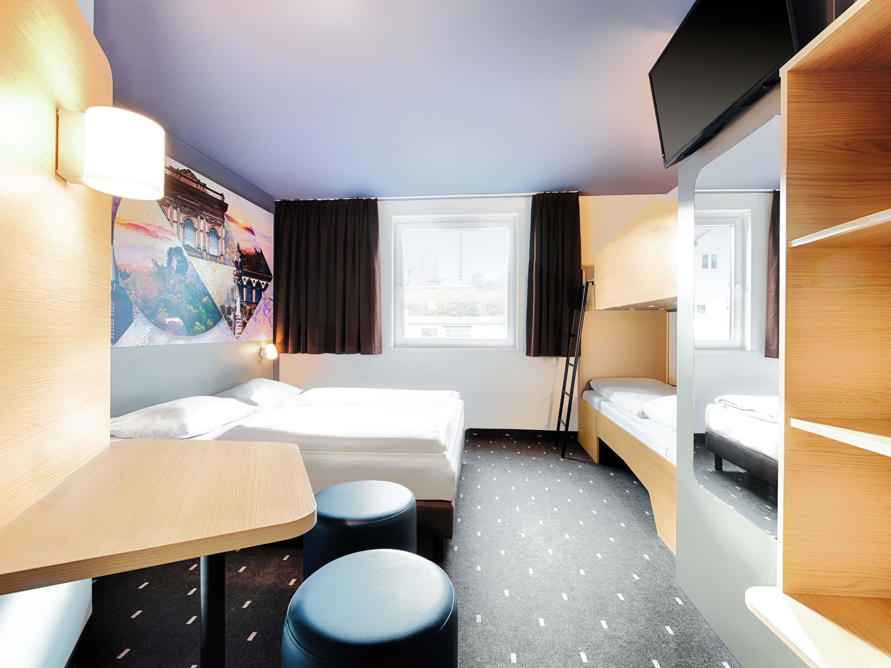 Une chambre d’hôtel de taille modeste, comprenant deux lits, une petite table, une chaise, un miroir et une fenêtre avec des rideaux.