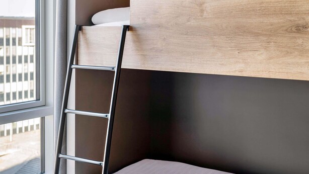 A bunk bed with a wooden headboard, a metal ladder, and a window with a view of buildings. - B&B Hotel Kaiserslautern (Kaiserslautern)