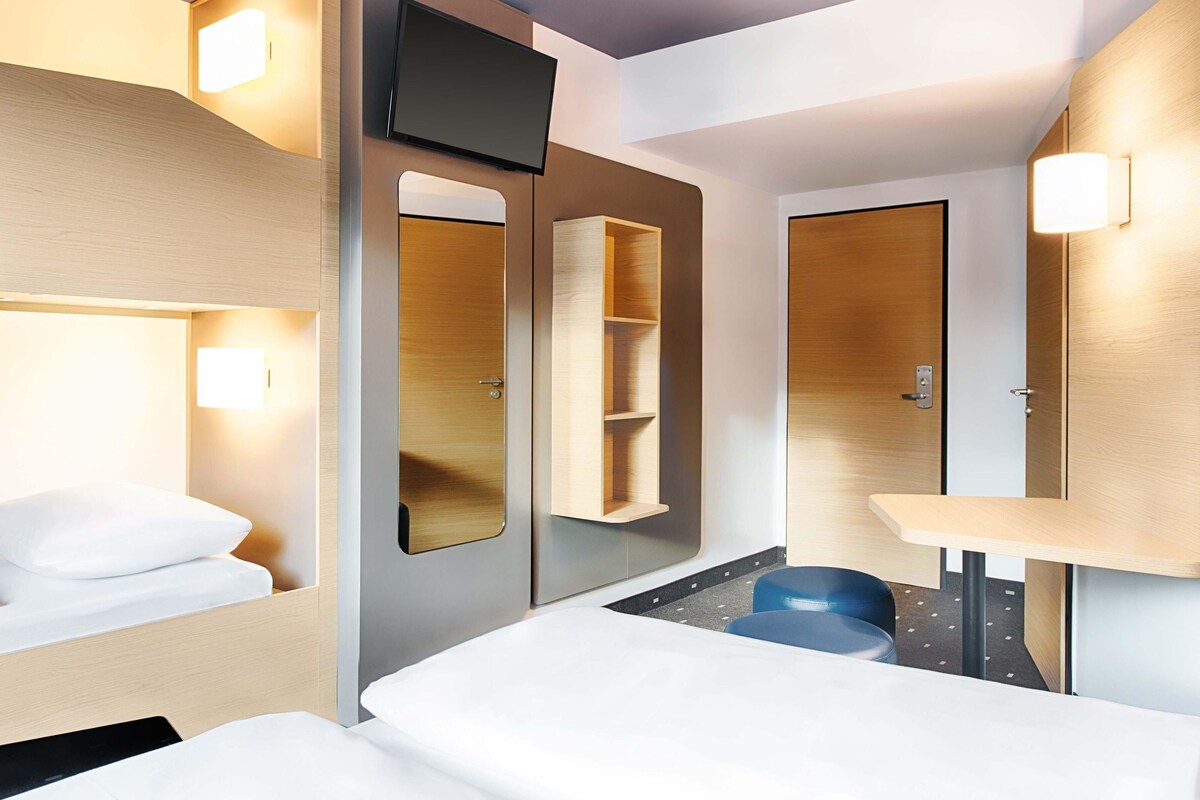 A modern hotel room with a bed, a desk, a chair, a TV, and built-in shelves.