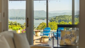 Une vue à travers une fenêtre donnant sur une terrasse, avec des chaises et une table, et offrant un panorama sur un lac et des montagnes.