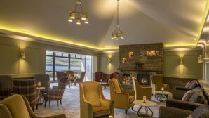 A spacious lounge area with comfortable seating, a stone fireplace, and large windows offering a view of the outdoors. - Dingle Skellig (Dingle)