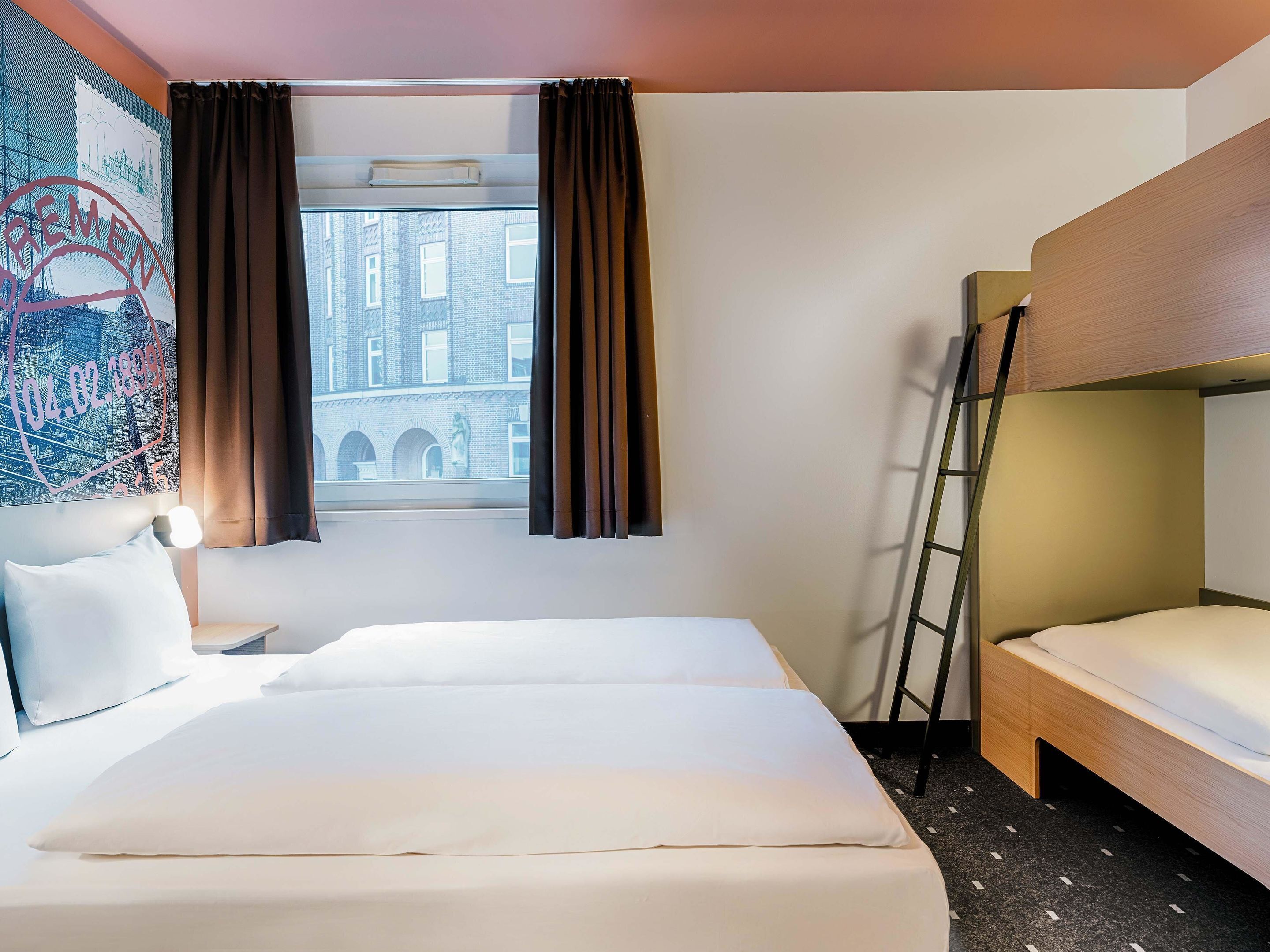 Una habitación de hotel con una cama grande, una cama más pequeña, una ventana con cortinas, una escalera y un cuadro de un paisaje urbano en la pared.