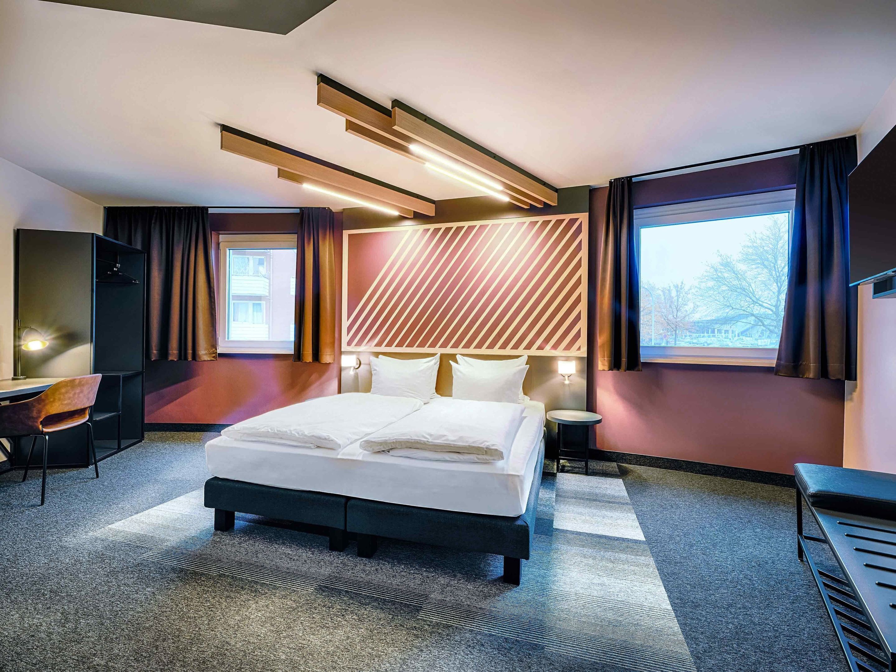 Ein modernes Hotelzimmer mit einem großen Bett, einem Schreibtisch und einem Stuhl. Es gibt ein Fenster mit Vorhängen, einen an der Wand befestigten Fernseher und einen Kopfteil mit einem geometrischen Muster.