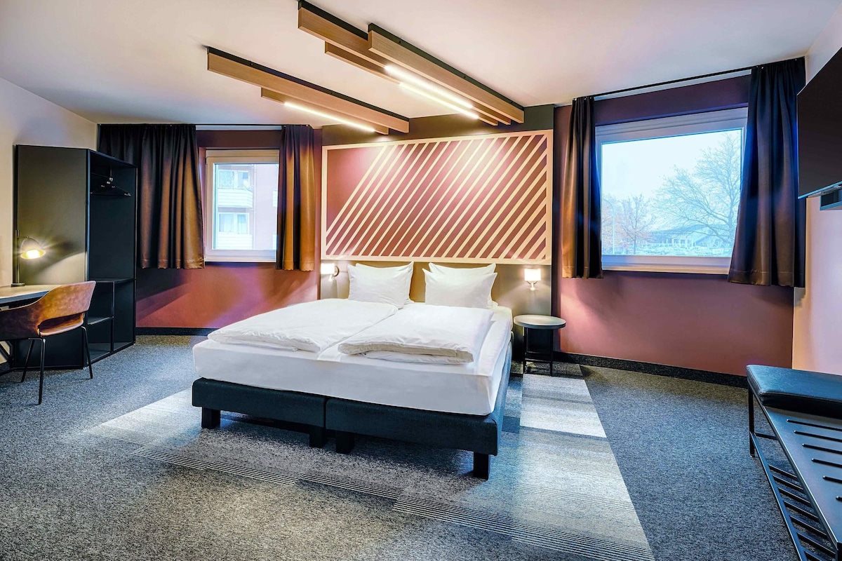Une chambre d’hôtel moderne avec un grand lit, un bureau et une chaise. On y trouve une fenêtre avec des rideaux, un téléviseur fixé au mur et une tête de lit ornée d’un motif géométrique.