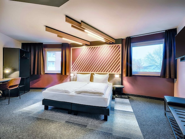 Une chambre d’hôtel moderne avec un grand lit, un bureau et une chaise. On y trouve une fenêtre avec des rideaux, un téléviseur fixé au mur et une tête de lit ornée d’un motif géométrique.