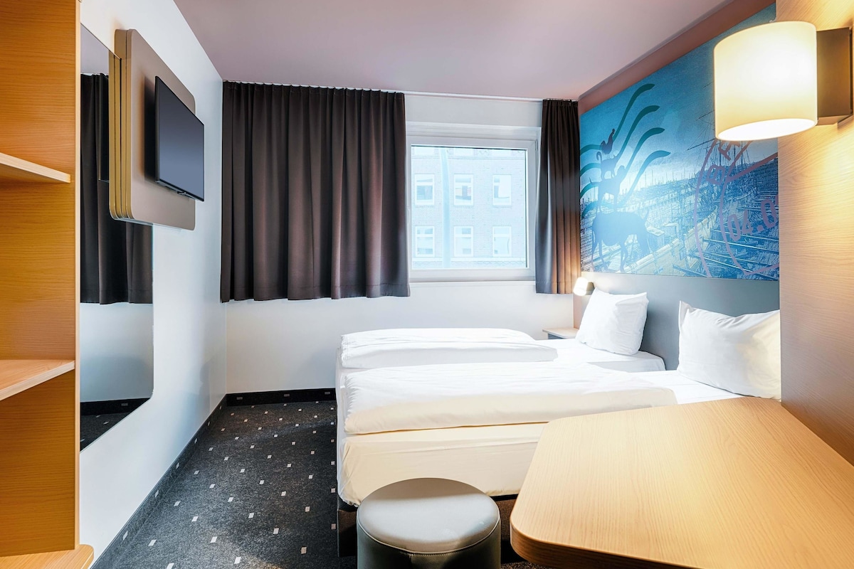 Une chambre d’hôtel avec un lit, un bureau, une chaise, une télévision et une fenêtre avec des rideaux.