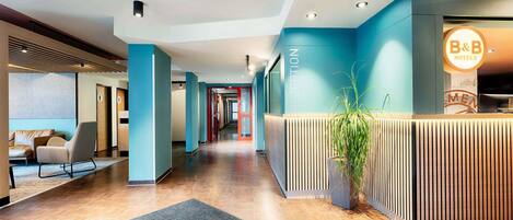 A modern hotel reception area with a reception desk, a seating area with a sofa, and a potted plant.
