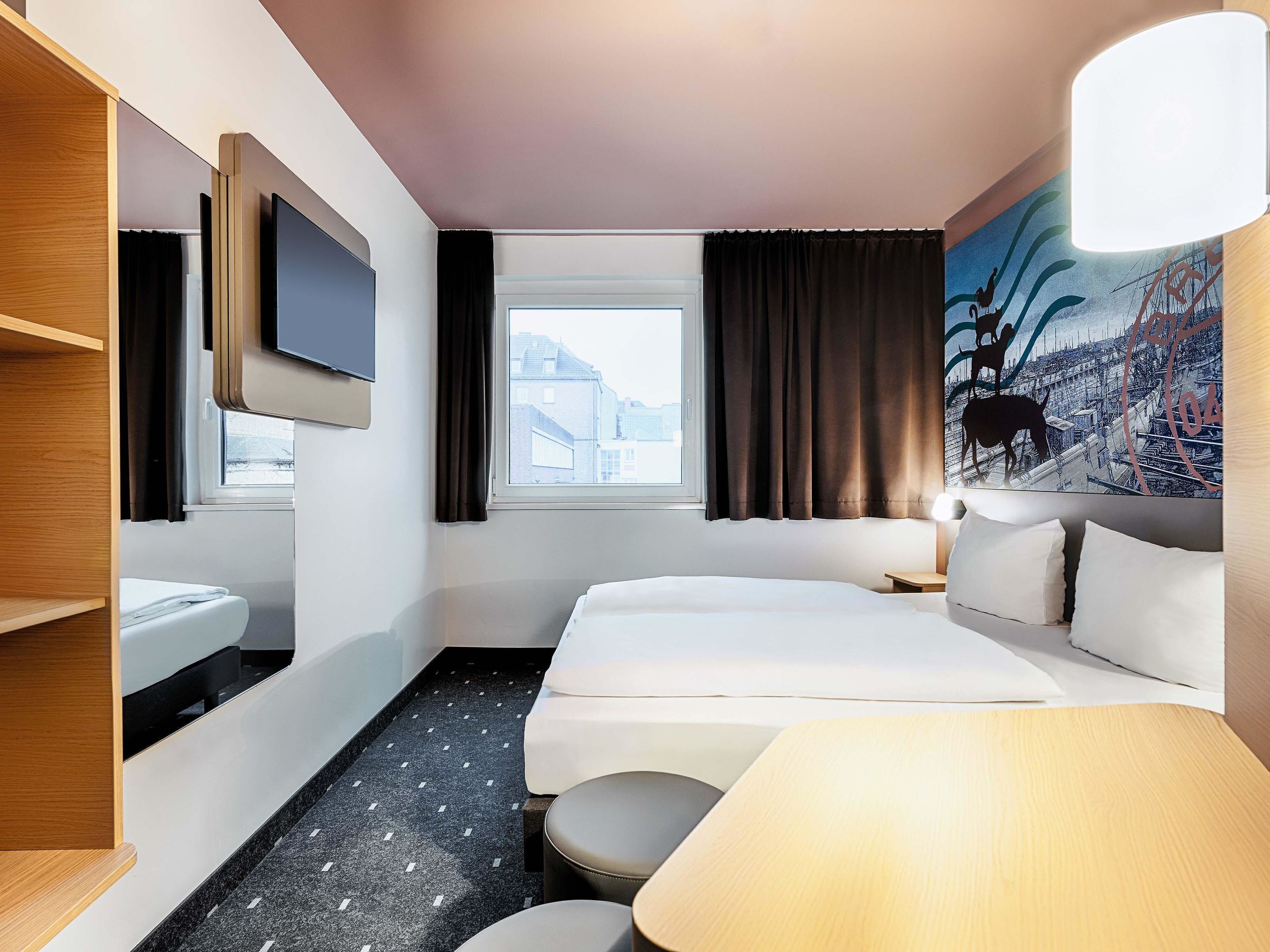 Ein Hotelzimmer mit Bett, Fernseher, Fenster mit Vorhängen, einem Gemälde und einer Lampe.