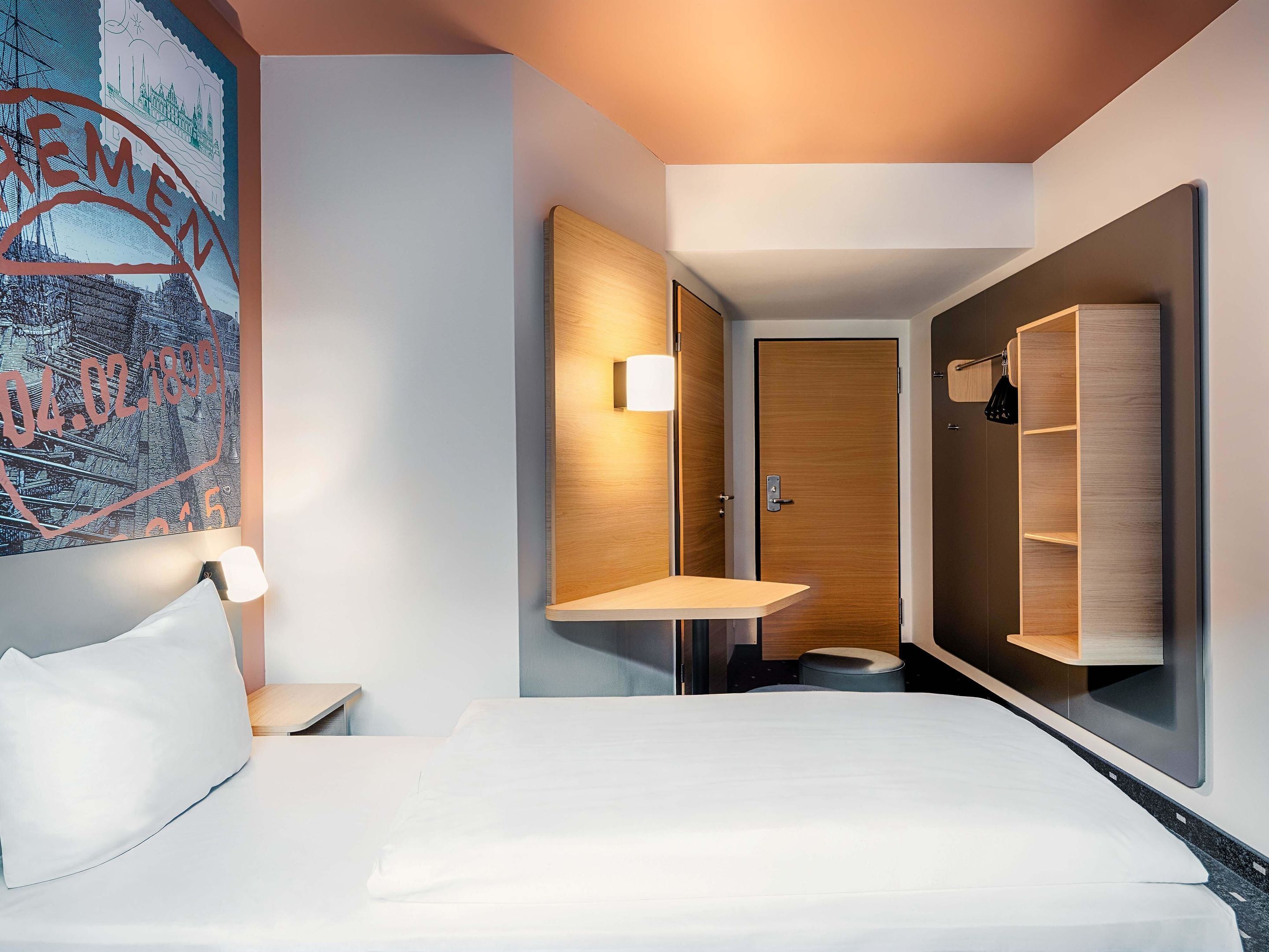 Una habitación de hotel moderna con una cama grande, una mesita de noche, un estante en la pared, un armario y un cuadro.