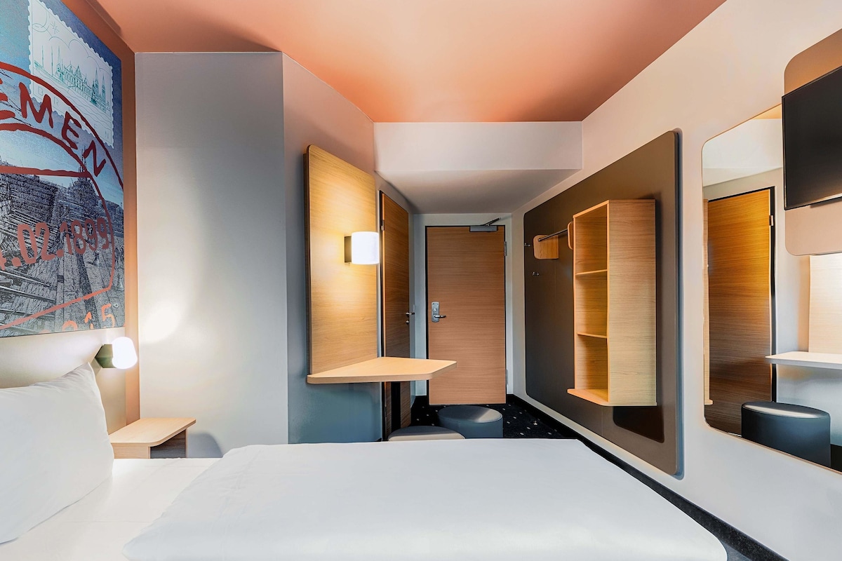 Une chambre d’hôtel moderne dotée d’un grand lit, d’étagères intégrées, d’une porte avec une poignée et d’un luminaire fixé au mur.