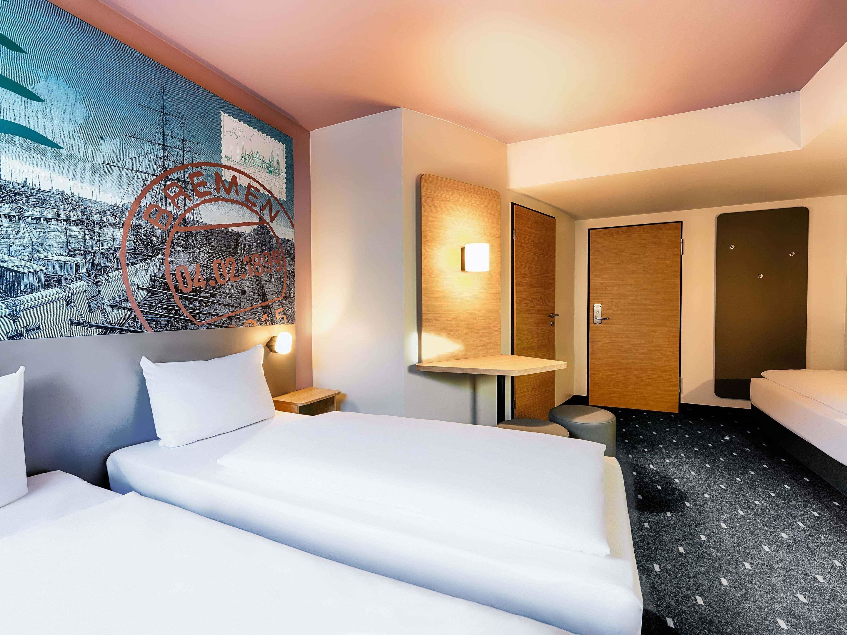 Habitación de hotel con una cama grande, una mesita de noche pequeña, una lámpara de pared, un espejo redondo, una puerta de madera y un piso alfombrado oscuro.
