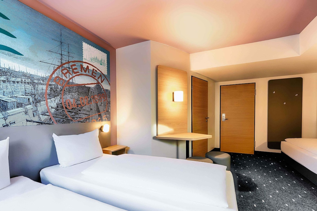 Une chambre d’hôtel avec un grand lit, une petite table de chevet, une commode, une lampe fixée au mur, un miroir rond, une porte en bois et un sol recouvert d’une moquette sombre.