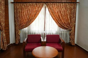 Ein Zimmer mit zwei roten Polstersesseln, einem Holztisch und einem Fenster mit gemusterten Vorhängen.